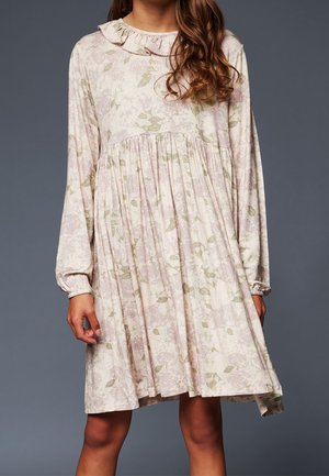 Femme portant une robe ample à manches longues et longueur genou, imprimée de fleurs, avec un col à volants, sur un fond uni sombre.