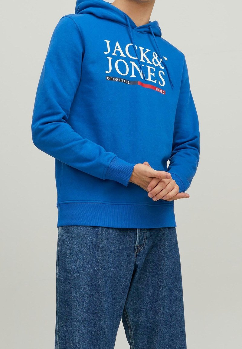 Muž v modré mikině Jack & Jones s bílým textem a modrými džíny, ruce složené před sebou, na světlém jednobarevném pozadí.