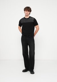 T-shirt preta com uma seção superior cinza escuro texturizada, combinada com jeans pretos de perna reta e sapatos pretos, num fundo branco.