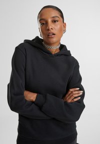 Femeie cu păr dat pe spate, purtând un hanorac negru, un lanț argintiu, cercei cerc și cu brațele încrucișate pe un fundal simplu.