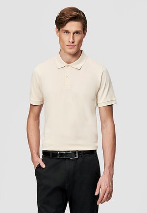 Mężczyzna w jasnobeżowej polówce z krótkim rękawem wsadzonej w czarne spodnie z czarnym paskiem, stojący na tle jednolitego szarego tła.