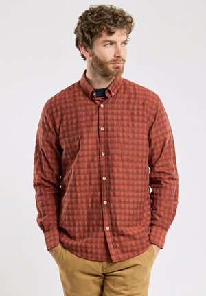 Chemise à manches longues à carreaux rouges avec un col boutonné, présentant un tissu texturé et une poche poitrine. Fermeture à boutons et ourlet arrondi.