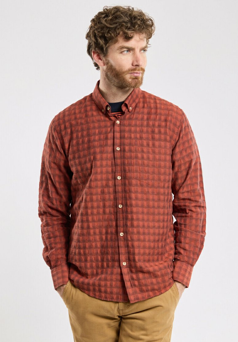 Chemise à manches longues à carreaux rouges avec un col boutonné, présentant un tissu texturé et une poche poitrine. Fermeture à boutons et ourlet arrondi.