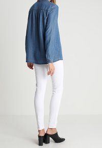 Camicia in denim blu medio con maniche lunghe, dettagli di pieghe sul retro e colletto con bottoni; abbinata a jeans skinny bianchi e mule nere con tacco.