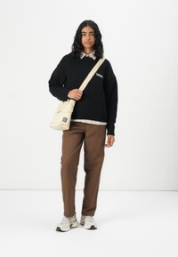 Zwart oversized sweater met wit logo, beige schoudertas, bruine broek en grijze sneakers. Model heeft lang, golvend haar en staat tegen een witte achtergrond.