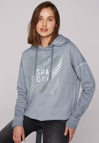 Felpa leggera blu con un design grafico argentato e la scritta "SPA DAY." Presenta un cappuccio con coulisse e maniche lunghe. Tessuto dalla texture morbida.