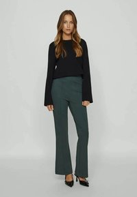 T-shirt preto de mangas longas com punhos largos combinado com calças verde-escuras de pernas largas. O conjunto tem uma textura suave e um design minimalista.