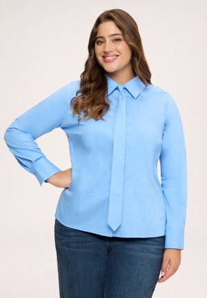 Smilende kvinde med mellemlangt brunt hår iført en lyseblå bluse med lange ærmer og bindedetalje samt mørkeblå jeans, hånd på hoften.