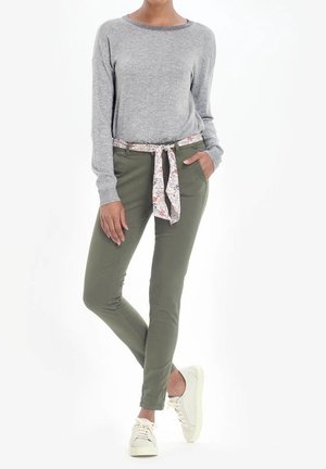 Person iført grå langærmet top, olivengrønne bukser med blomstret stofbælte, hvide sneakers, stående med den ene hånd i lommen og benene krydset.