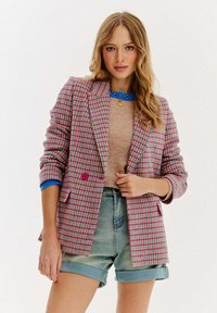 Mladá žena v barevném kostkovaném saku, béžovém svetru, modrém topu, džínových šortkách a zlatém náhrdelníku, stojící na bílém pozadí.