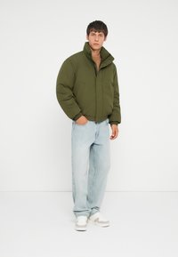 Olive green puffer jacket, relaxed fit, high collar, front pockets. Light blue jeans, casual style, paired with white sneakers.