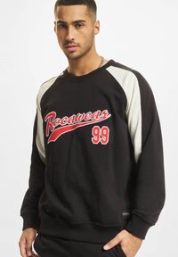 Svart hettegenser med kremfargede raglanermer, med rød "Rocawear" logo og "99" på brystet. Mykt materiale med ribbestrikkede mansjetter og nederkant.