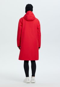 Impermeabile rosso con cappuccio, lunghezza al ginocchio, texture liscia, maniche lunghe e polsini regolabili. Indossato sopra leggings neri e sneakers bianche.