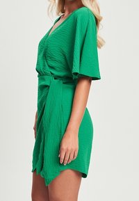 Vihreä kietaisumekko, jossa on V-pääntie, lyhyet lepattelevat hihat, teksturoitu kangas ja vyötärölle sidottava nauha säädettävää istuvuutta varten.