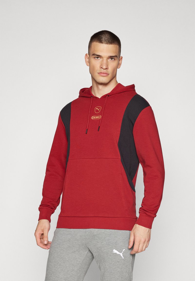 Felpa rossa con pannelli laterali neri, cappuccio regolabile e logo dorato sul petto. Abbinata a pantaloni della tuta grigi con logo bianco.