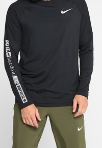 Mustapituinen urheilupaita pitkällä hihalla, jossa on valkoinen Nike-logo rinnan alueella ja "Just Do It" -teksti vasemmassa hihassa. Esteettisesti sileä kangas, normaali istuvuus.