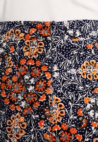 Gros plan sur un tissu avec un fond bleu foncé, un motif floral orange et blanc, et une fermeture éclair visible au centre en haut.