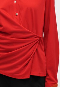 Rote Bluse mit langen Ärmeln, goldenen Knöpfen und einem gerafften Drehdetail an der Taille, getragen zu schwarzen Hosen.