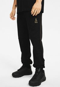 Mężczyzna w czarnych dresach z złotym logo Dakar i czarnych masywnych sneakersach, stojący na białym tle.