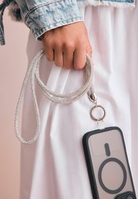 Hånd, der holder en glitrende perlerem, der er fastgjort til en smartphone med sort etui, iført en hvid nederdel og en denimjakkeærm.