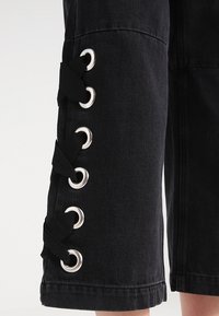 Svart denimplagg med öljett hårdvara och snörningsdetaljer längs sidsömmen, med rak skärning och strukturerad tyg.