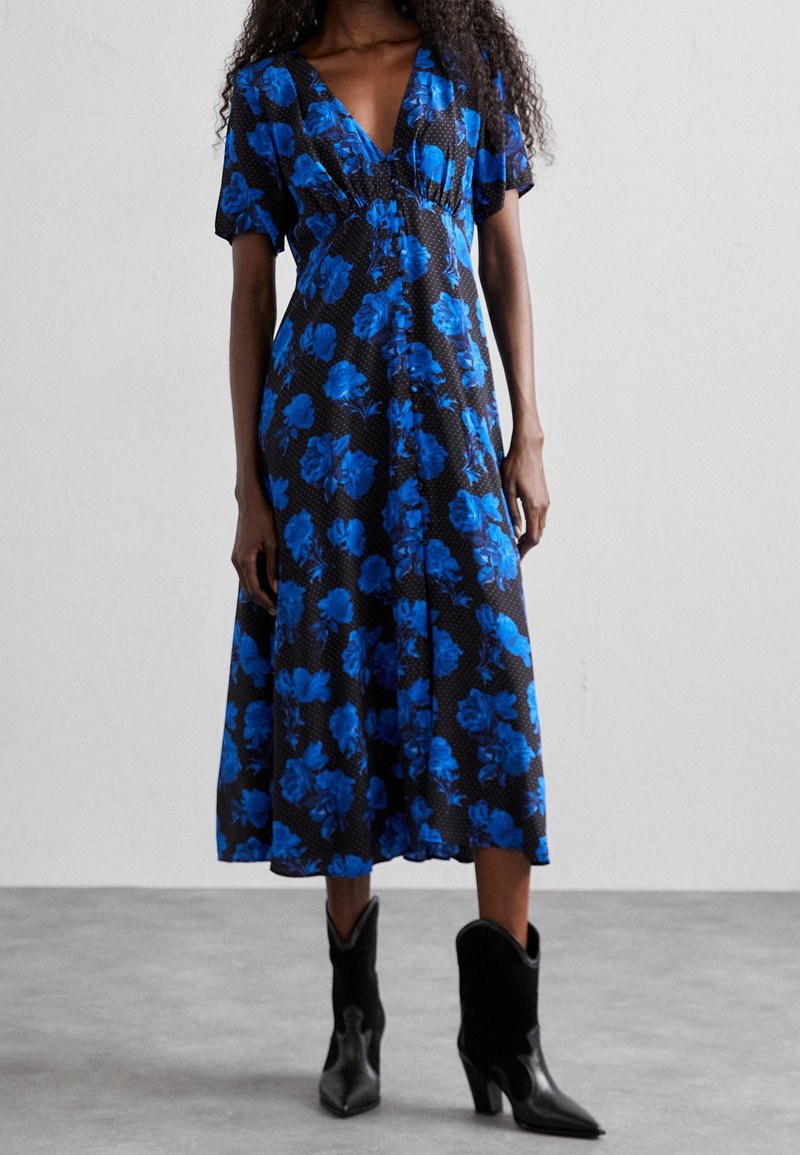 Femme portant une robe midi noire à imprimé floral bleu vif et manches courtes, associée à des bottines noires à talons.