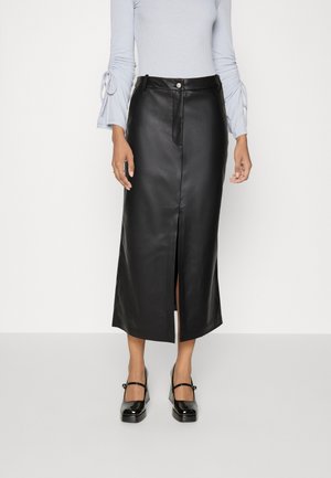 Vrouw draagt een zwarte leren midi-rok met een voorsplit, lichtgrijze top met lange mouwen en zwarte lak Mary Jane hakken.