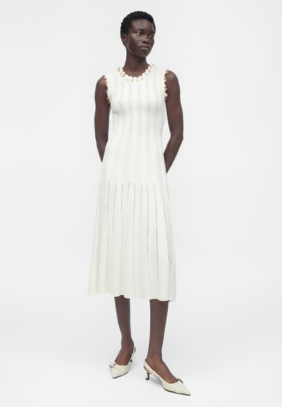 Γυναίκα που στέκεται φορώντας ένα λευκό πλεκτό μίντι φόρεμα χωρίς μανίκια με floral τελείωμα και κρεμ μυτερά παπούτσια με χαμηλό τακούνι.