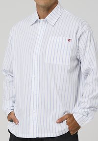 Λευκό πουκάμισο με στενές μπλε ρίγες, μακριά μανίκια, τσέπη στο στήθος και μικρό κόκκινο λογότυπο στην αριστερή πλευρά. Μαλακή υφή υφάσματος.