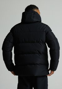 Svart puffjakke med strammet hette, quiltet design og elastiske mansjetter. Har en glatt, glinsende tekstur og en avslappet passform.