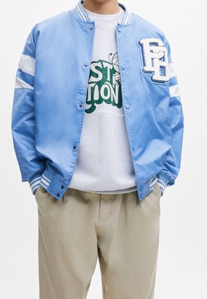 Mand iført lyseblå varsityjakke med hvide striber og beige bukser, hænderne i lommerne, delvist synlig grøn grafisk sweatshirt under.