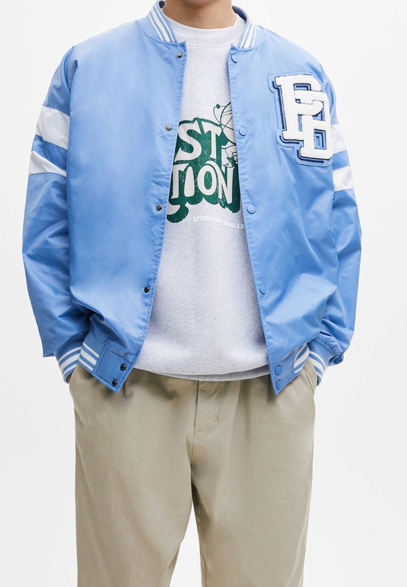 Homme portant une veste varsity bleu clair avec des bandes blanches et un pantalon beige, mains dans les poches, montrant partiellement un sweat-shirt graphique vert en dessous.
