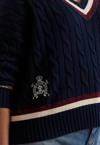 Temno modri pulover z vzorcem kabela in bordo ter belimi črtami na spodnjem robu in izrezu, z vezenim logotipom na levi strani, nošen z modrimi kavbojkami.