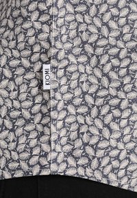 Camicia a maniche corte in tessuto blu navy con motivi di foglie bianche. Include una piccola etichetta bianca con la scritta "KOMI" vicino all'orlo.