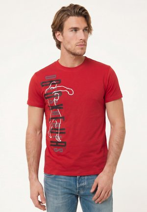 Homme portant un t-shirt rouge à manches courtes avec un texte noir vertical et un design en silhouette blanche, associé à un jean bleu, debout sur fond blanc.
