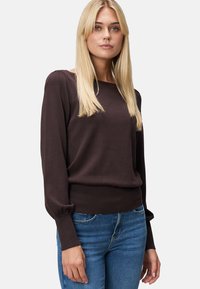 Femme blonde portant un pull long à manches longues marron foncé avec des poignets bouffants et un jean bleu, posant devant un fond clair.