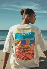 Bela oversized majica s pisanim grafičnim prikazom plaže s cvetličnimi in črtastimi stoli, pogled od zadaj, ozadje peščena plaža.