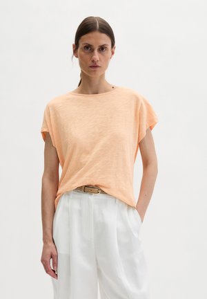 Kobieta o brązowych włosach związanych do tyłu, ubrana w luźny, brzoskwiniowy top z krótkim rękawem, włożony w białe spodnie z wysokim stanem i beżowym paskiem.