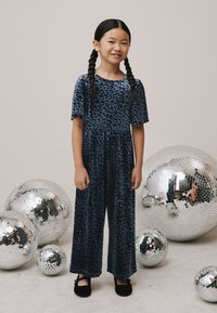 Tumši zils samta kombinezons ar leoparda rakstu, īsām piedurknēm un platiem galiem, sapārots ar melniem apaviem, fonā sudraba disko bumbas.