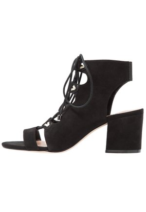 Sort ruskind højhælede sandal med åben tå, snørelukning foran og blokhæl. Har udskæringsdesign og sølvfarvede detaljer.