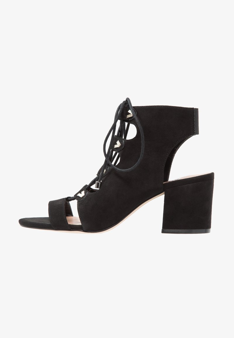 Sandale à talon haut en daim noir avec bout ouvert, lacets à l'avant et talon bloc. Présente un design ajouré et des accents argentés.