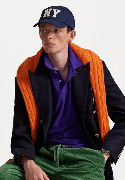 Blazer navy com botões dourados, suéter laranja de malha tricotada colocado sobre os ombros, camisa polo roxa, calças de corduroy verde, boné navy da NY.