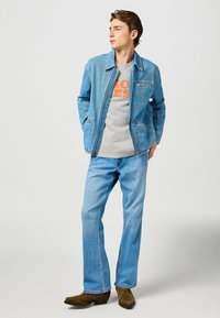 Chaqueta de mezclilla en azul claro con diseño de cuello, combinada con una camiseta gris que tiene texto naranja, y jeans rectos azul. Botas marrones.