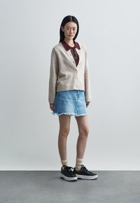 Beige wollen vest met knopen, daaronder een donkerrode blouse met kraag, gecombineerd met een lichte denim rok met rafelige zoom en zwarte sneakers.