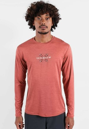 Homme aux cheveux bouclés portant une chemise à manches longues couleur rouille avec le logo "OXBOW", debout devant un fond clair uni.