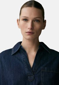 Chemise en denim de couleur bleu foncé, avec un col pointu et un patte de boutonnage partielle. Présente une poche sur la poitrine et des détails de couture prononcés.