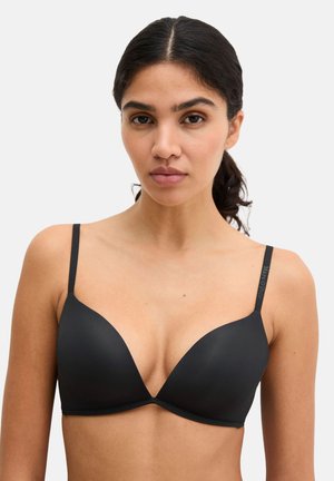 Młoda kobieta o ciemnych włosach związanych w kucyk, ubrana w prosty czarny usztywniany biustonosz z cienkimi regulowanymi ramiączkami.