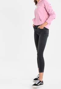 Jeans neri aderenti abbinati a una felpa rosa con tasca frontale e coulisse. Il modello indossa sneakers nere e bianche.