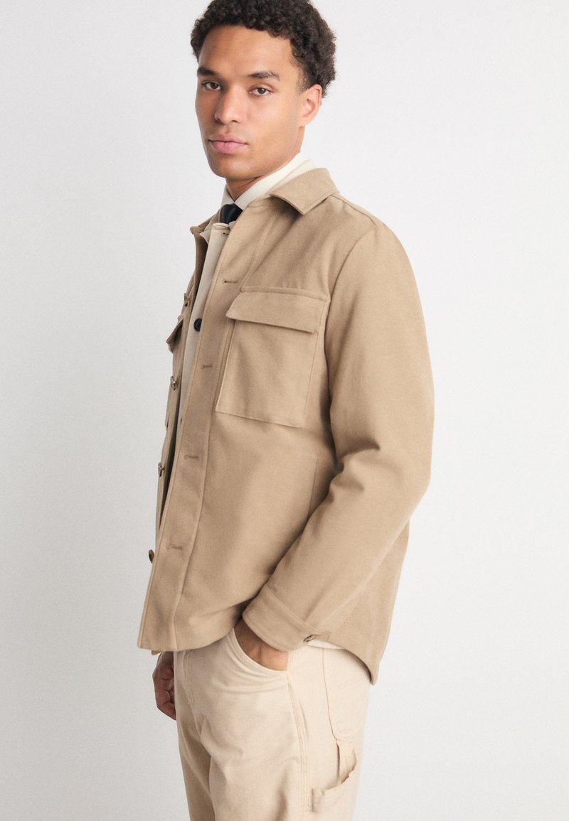 Veste beige avec un devant à boutons, deux poches poitrine et un col. Fabriquée en tissu doux, elle présente une coupe décontractée et un design minimal.