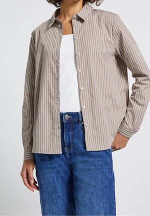 Persona que lleva una camisa beige y blanca de rayas verticales sobre una camiseta blanca con vaqueros azules, mano apoyada en la cadera.